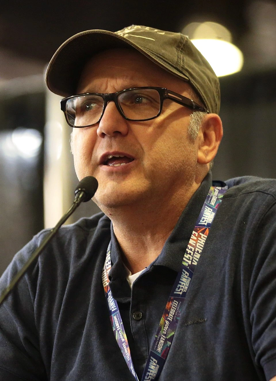 Portrait photo of Paris Themmen speaking at a microphone, wearing glasses and a cap.