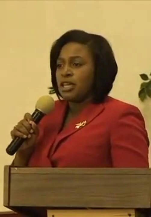 Lovely A. Warren speaking at a podium in a red jacket, holding a microphone